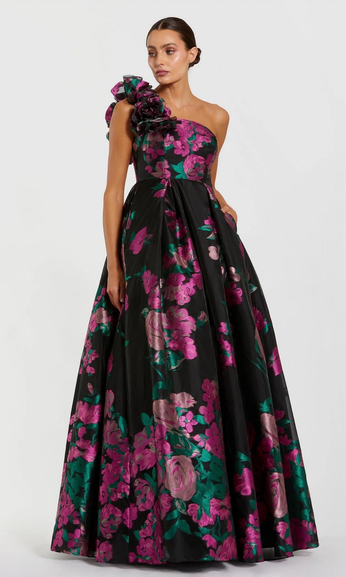 A woman wears the Mac Duggal 11967 long formal dress in Black Multi—a floor-length, one-shoulder ballgown featuring pink and green floral prints and a dramatic shoulder ruffle.